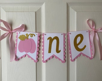 May include: A pink and gold "one" banner with a pumpkin design. The banner has three pennant-shaped flags with scalloped edges. The word "one" is in gold glitter letters. Pink ribbons are tied on each end.