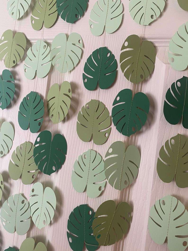 K&ouml;nnte beinhalten: Eine Girlande aus Papier-Monstera-Bl&auml;ttern in verschiedenen Gr&uuml;nt&ouml;nen, auf d&uuml;nnen Schn&uuml;ren aufgereiht. Die Bl&auml;tter haben ein detailliertes, ausgeschnittenes Design und sind vor einem hellen Hintergrund angeordnet, wodurch ein tropisches, dekoratives Thema entsteht.