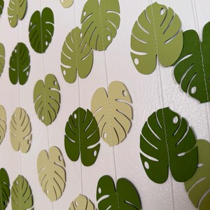 Puede incluir: Guirnalda de hojas de papel en tonos verdes y crema. La decoraci&oacute;n de tem&aacute;tica tropical presenta hojas de monstera recortadas, ensartadas en un hilo transparente. Las hojas var&iacute;an en color y est&aacute;n dispuestas sobre un fondo blanco.