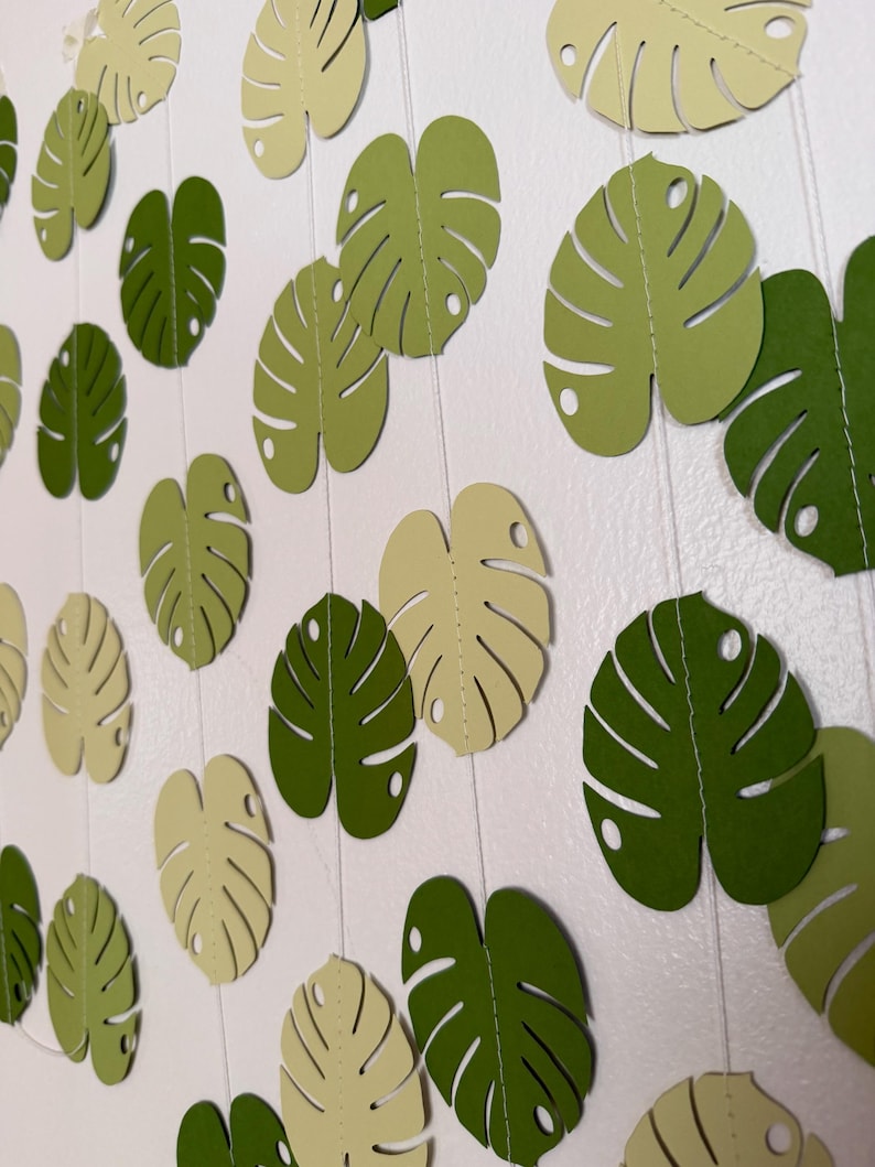 Puede incluir: Guirnalda de hojas de papel en tonos verdes y crema. La decoraci&oacute;n de tem&aacute;tica tropical presenta hojas de monstera recortadas, ensartadas en un hilo transparente. Las hojas var&iacute;an en color y est&aacute;n dispuestas sobre un fondo blanco.