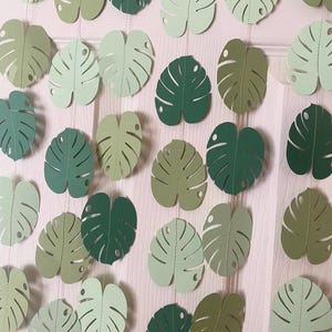 Puede incluir: Una guirnalda de hojas de Monstera de papel en varios tonos de verde. Las hojas est&aacute;n cortadas con patrones de venas detallados y ensartadas. El texto "Style 2" es visible en la parte inferior.
