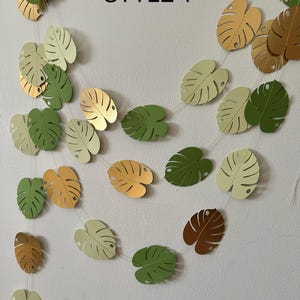 Puede incluir: Una guirnalda de hojas de papel en varios tonos de verde, dorado y marr&oacute;n, ensartadas. El texto "STYLE 1" se muestra en negro. Las hojas tienen forma de hoja de monstera.