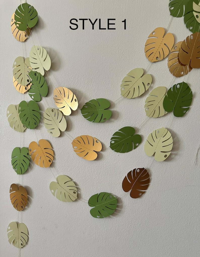 Puede incluir: Una guirnalda de hojas de papel en varios tonos de verde, dorado y marr&oacute;n, ensartadas. El texto "STYLE 1" se muestra en negro. Las hojas tienen forma de hoja de monstera.