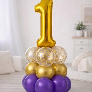 May include: A balloon arrangement for a first birthday celebration. The display features a large gold foil number one balloon atop a base of purple, gold, and confetti-filled balloons. The arrangement is set against a soft, neutral background.