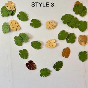 Puede incluir: Una guirnalda de hojas de papel en tonos verdes, amarillos y marrones, ensartadas con un hilo fino y transparente. El texto "STYLE 3" est&aacute; impreso en negro en la parte superior. Las hojas tienen un dise&ntilde;o de hoja de monstera.