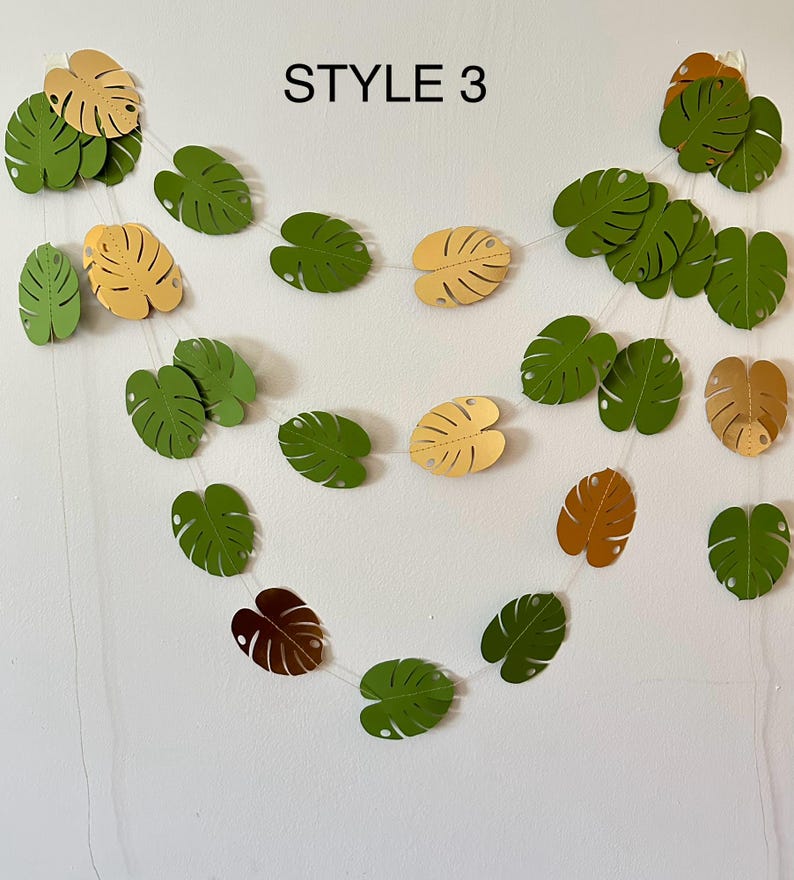 Puede incluir: Una guirnalda de hojas de papel en tonos verdes, amarillos y marrones, ensartadas con un hilo fino y transparente. El texto "STYLE 3" est&aacute; impreso en negro en la parte superior. Las hojas tienen un dise&ntilde;o de hoja de monstera.
