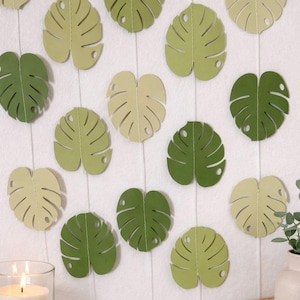 May include: Decorative paper Monstera leaves in shades of green and cream are arranged on a white background. The leaves are cut with detailed vein patterns and a hole near the stem. A lit candle and a small plant are visible.