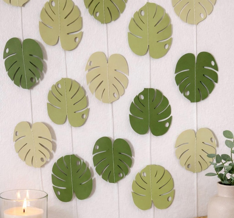 K&ouml;nnte beinhalten: Dekorative Monstera-Bl&auml;tter aus Papier in Gr&uuml;n- und Cremet&ouml;nen, angeordnet auf einem wei&szlig;en Hintergrund. Die Bl&auml;tter sind mit detaillierten Adern und einem Loch am Stiel versehen. Eine brennende Kerze und eine kleine Pflanze sind ebenfalls zu sehen.