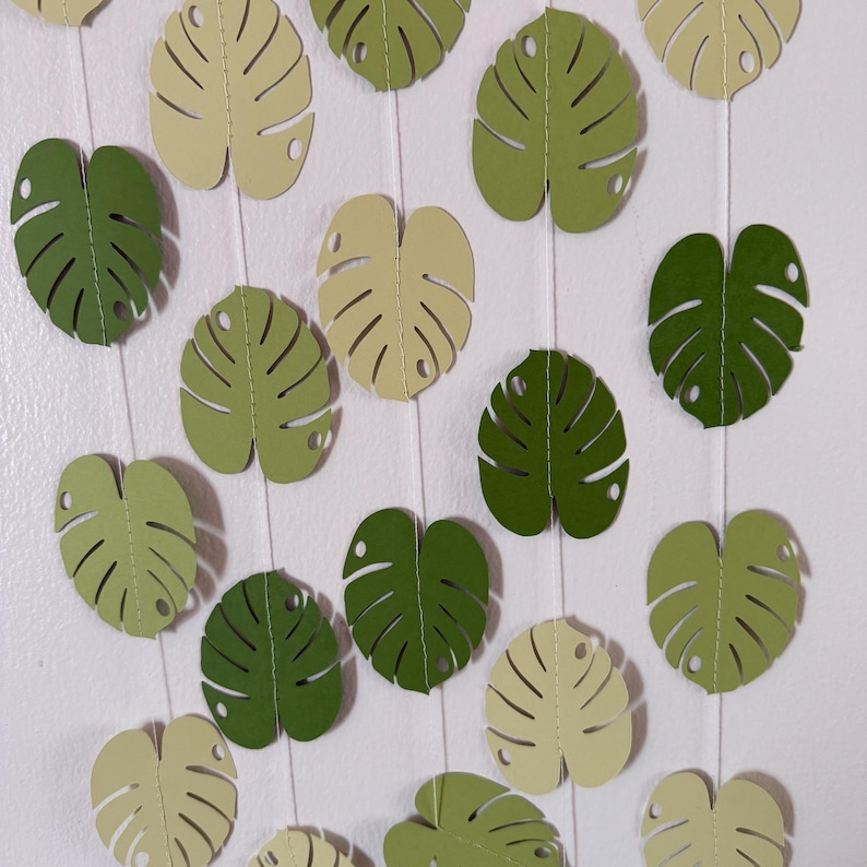 Puede incluir: Una guirnalda de hojas de monstera de papel en varios tonos de verde y crema. Las hojas est&aacute;n ensartadas en una cuerda blanca, creando un tema tropical y bot&aacute;nico. Las hojas tienen detalles recortados y est&aacute;n dispuestas sobre un fondo blanco.