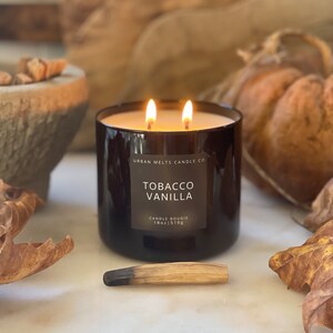 Puede incluir: Una vela de vidrio negro con una etiqueta que dice "Urban Melts Candle Co. Tobacco Vanilla Candle Bougie 18oz | 510g". La vela est&aacute; encendida y tiene dos mechas.