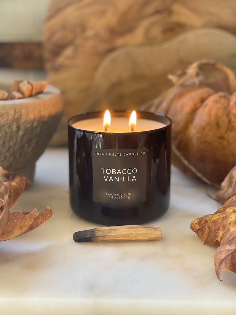 Puede incluir: Una vela de vidrio negro con una etiqueta que dice "Urban Melts Candle Co. Tobacco Vanilla Candle Bougie 18oz | 510g". La vela est&aacute; encendida y tiene dos mechas.