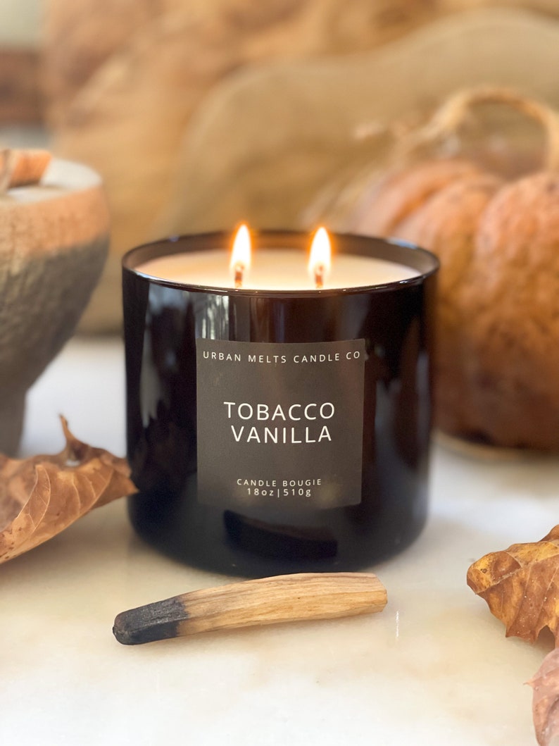 Puede incluir: Un tarro de vela de vidrio negro con una etiqueta blanca que dice "URBAN MELTS CANDLE CO. TOBACCO VANILLA CANDLE BOUGIE 18oz|510g". La vela est&aacute; encendida y tiene dos mechas.