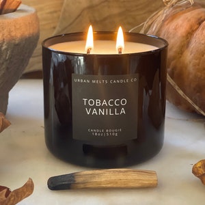 Puede incluir: Un tarro de vela de vidrio negro con dos mechas, etiquetado como "Tobacco Vanilla" por Urban Melts Candle Co. La vela es de 18 oz o 510g.