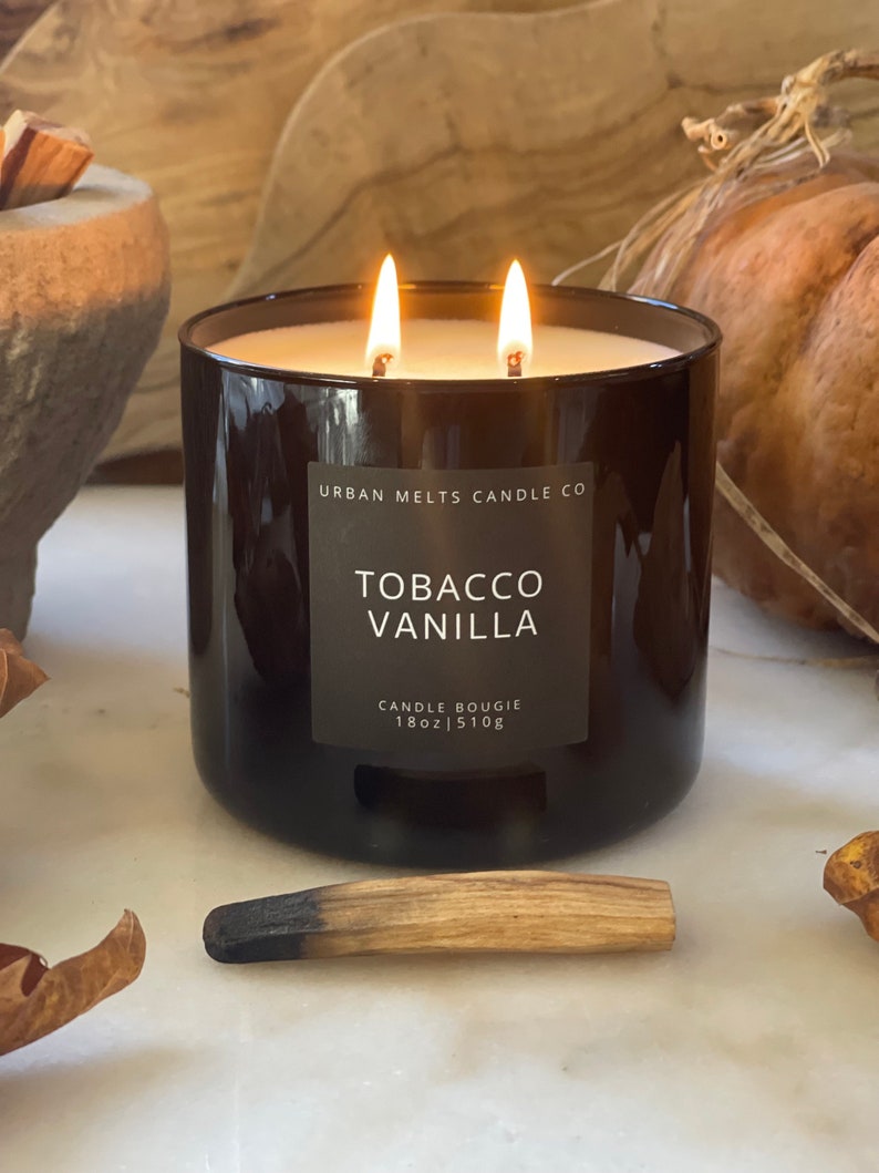 Puede incluir: Un tarro de vela de vidrio negro con dos mechas, etiquetado como "Tobacco Vanilla" por Urban Melts Candle Co. La vela es de 18 oz o 510g.