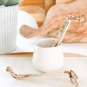 May include: Two gold candle wick trimmers, one with a decorative flower design, in a white ceramic cup on a white marble surface.