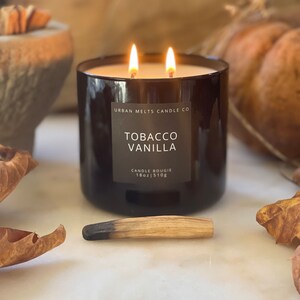 Puede incluir: Un tarro de vela de vidrio negro con una etiqueta marr&oacute;n que dice "Urban Melts Candle Co. Tobacco Vanilla Candle Bougie 18oz | 510g". La vela est&aacute; encendida y tiene dos mechas. La vela est&aacute; sentada sobre una superficie blanca con una cerilla de madera y hojas de oto&ntilde;o.