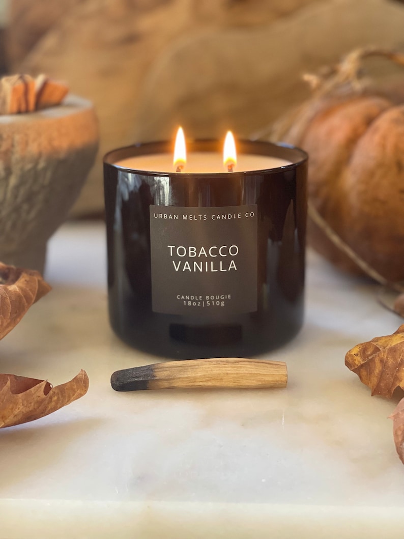 Puede incluir: Un tarro de vela de vidrio negro con una etiqueta marr&oacute;n que dice "Urban Melts Candle Co. Tobacco Vanilla Candle Bougie 18oz | 510g". La vela est&aacute; encendida y tiene dos mechas. La vela est&aacute; sentada sobre una superficie blanca con una cerilla de madera y hojas de oto&ntilde;o.