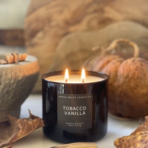 Puede incluir: Una vela de vidrio negro con dos mechas, etiquetada "Tobacco Vanilla" por Urban Melts Candle Co. La vela es de 18 oz / 510g y est&aacute; sobre una superficie blanca con hojas de oto&ntilde;o y una cerilla de madera.