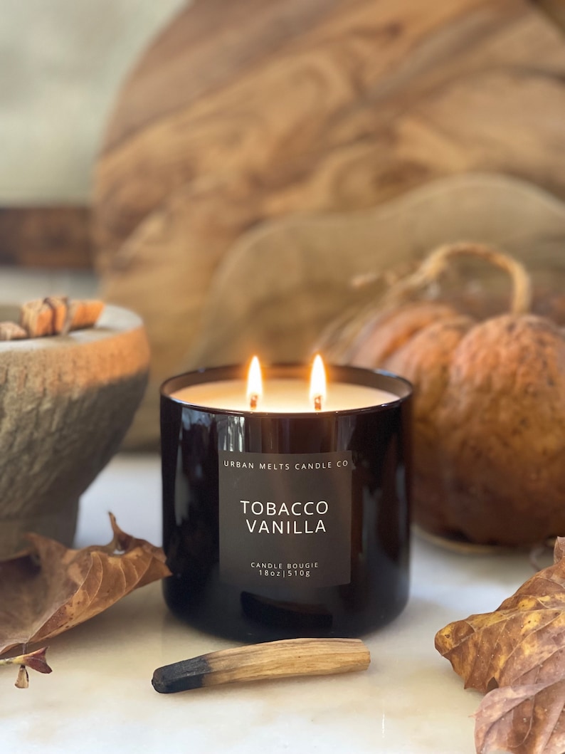 Puede incluir: Una vela de vidrio negro con dos mechas, etiquetada "Tobacco Vanilla" por Urban Melts Candle Co. La vela es de 18 oz / 510g y est&aacute; sobre una superficie blanca con hojas de oto&ntilde;o y una cerilla de madera.