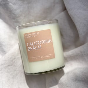 May include: A clear glass candle jar with a white soy wax candle inside. The label is peach colored with white text that reads "Urban Melts Candle Co. California Beach Hand Poured in California 11oz | Soy Wax Candle".