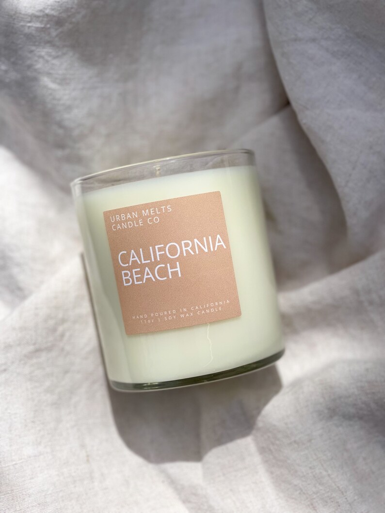 May include: A clear glass candle jar with a white soy wax candle inside. The label is peach colored with white text that reads "Urban Melts Candle Co. California Beach Hand Poured in California 11oz | Soy Wax Candle".