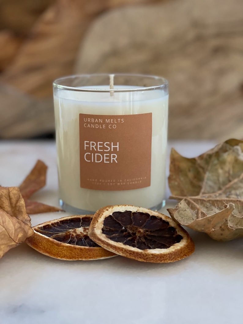 May include: A clear glass candle jar with a white soy wax candle inside. The candle is labeled "Urban Melts Candle Co. Fresh Cider" with a brown label. The candle is sitting on a white surface with dried orange slices and fall leaves.