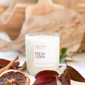 May include: A clear glass candle jar with a white label that reads "URBAN MELTS CANDLE CO FRESH CIDER HAND POURED IN CALIFORNIA 11oz | SOY WAX CANDLE". The candle is surrounded by cinnamon sticks and a dried orange slice.