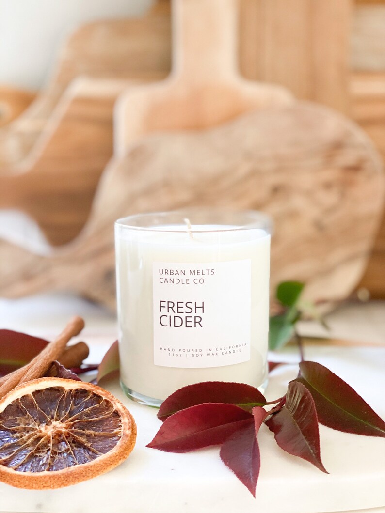 May include: A clear glass candle jar with a white label that reads "URBAN MELTS CANDLE CO FRESH CIDER HAND POURED IN CALIFORNIA 11oz | SOY WAX CANDLE". The candle is surrounded by cinnamon sticks and a dried orange slice.