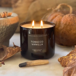 Puede incluir: Una vela de vidrio negro con una etiqueta blanca que dice "Urban Melts Candle Co. Tobacco Vanilla Candle Bougie 18oz | 510g". La vela est&aacute; encendida y tiene dos mechas.