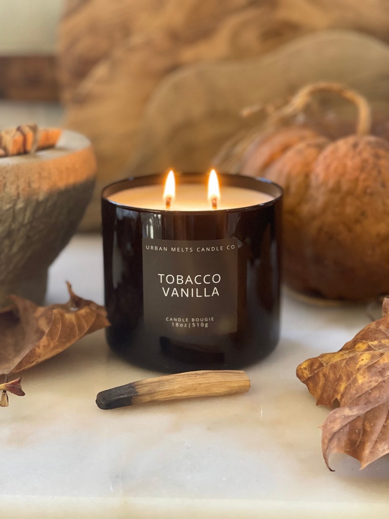 Puede incluir: Una vela de vidrio negro con una etiqueta blanca que dice "Urban Melts Candle Co. Tobacco Vanilla Candle Bougie 18oz | 510g". La vela est&aacute; encendida y tiene dos mechas.