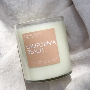 May include: A clear glass candle jar with a beige label that reads "URBAN MELTS CANDLE CO CALIFORNIA BEACH HAND POURED IN CALIFORNIA 11 OZ SOY WAX CANDLE".