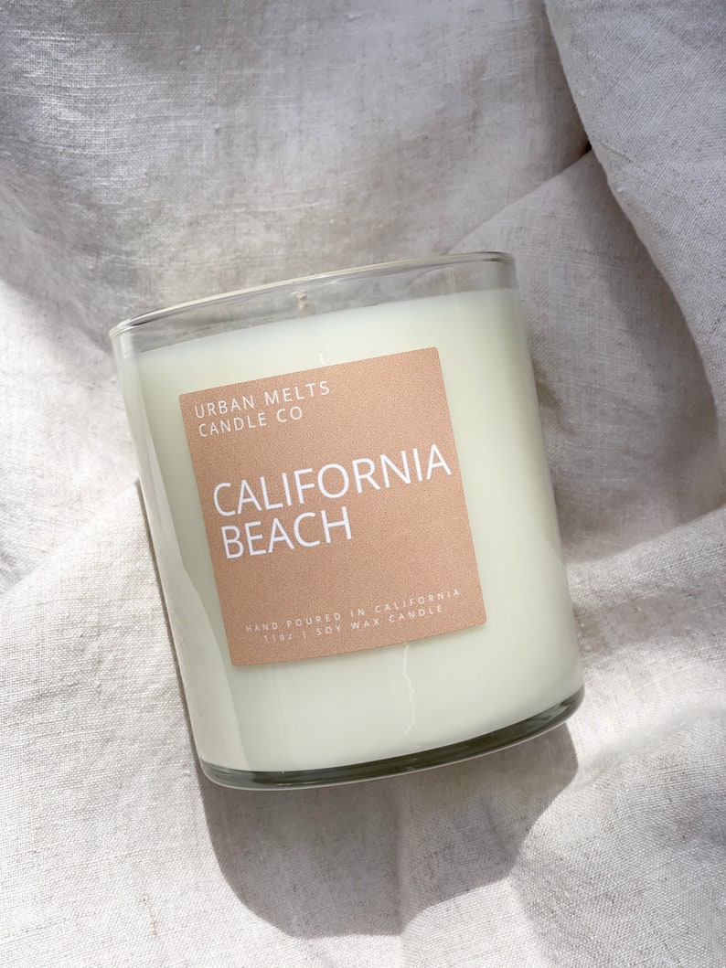 May include: A clear glass candle jar with a beige label that reads "URBAN MELTS CANDLE CO CALIFORNIA BEACH HAND POURED IN CALIFORNIA 11 OZ SOY WAX CANDLE".