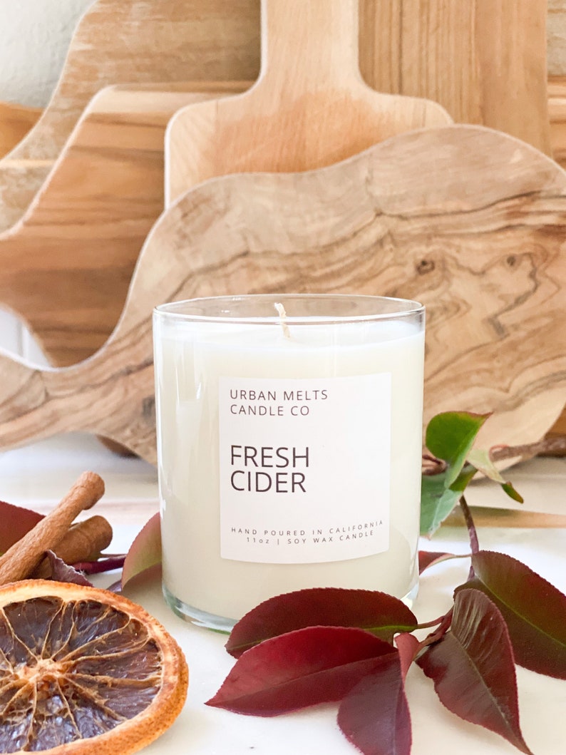 May include: A clear glass candle jar with a white label that reads "URBAN MELTS CANDLE CO FRESH CIDER HAND POURED IN CALIFORNIA 11oz | SOY WAX CANDLE". The candle is surrounded by cinnamon sticks, dried orange slices, and red leaves.