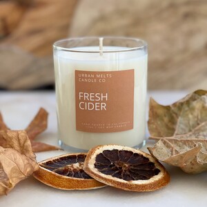 May include: A clear glass candle jar with a white candle inside. The label on the jar reads "Urban Melts Candle Co. Fresh Cider" and "Hand Poured in California. 100% Soy Wax Candle". The candle is surrounded by dried orange slices and fall leaves.