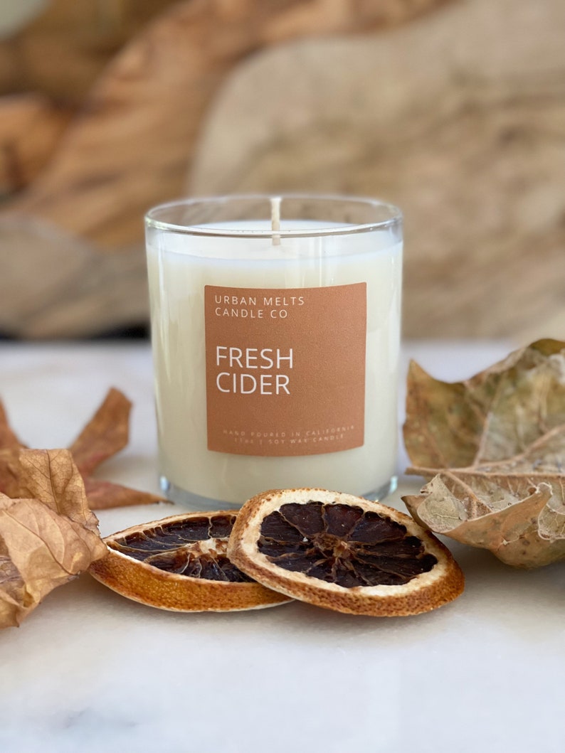 May include: A clear glass candle jar with a white candle inside. The label on the jar reads "Urban Melts Candle Co. Fresh Cider" and "Hand Poured in California. 100% Soy Wax Candle". The candle is surrounded by dried orange slices and fall leaves.