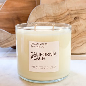 May include: A clear glass candle jar with a white label that reads "URBAN MELTS CANDLE CO CALIFORNIA BEACH HAND POURED IN CALIFORNIA 18oz | SOY WAX CANDLE".