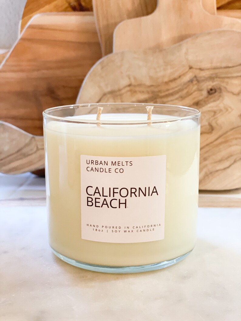 May include: A clear glass candle jar with a white label that reads "URBAN MELTS CANDLE CO CALIFORNIA BEACH HAND POURED IN CALIFORNIA 18oz | SOY WAX CANDLE".