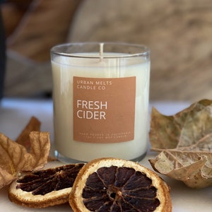 May include: A clear glass candle jar with a brown label that reads "Urban Melts Candle Co. Fresh Cider" and the text "Hand Poured in California 100% Soy Wax Candle". The candle is surrounded by dried orange slices and fall leaves.