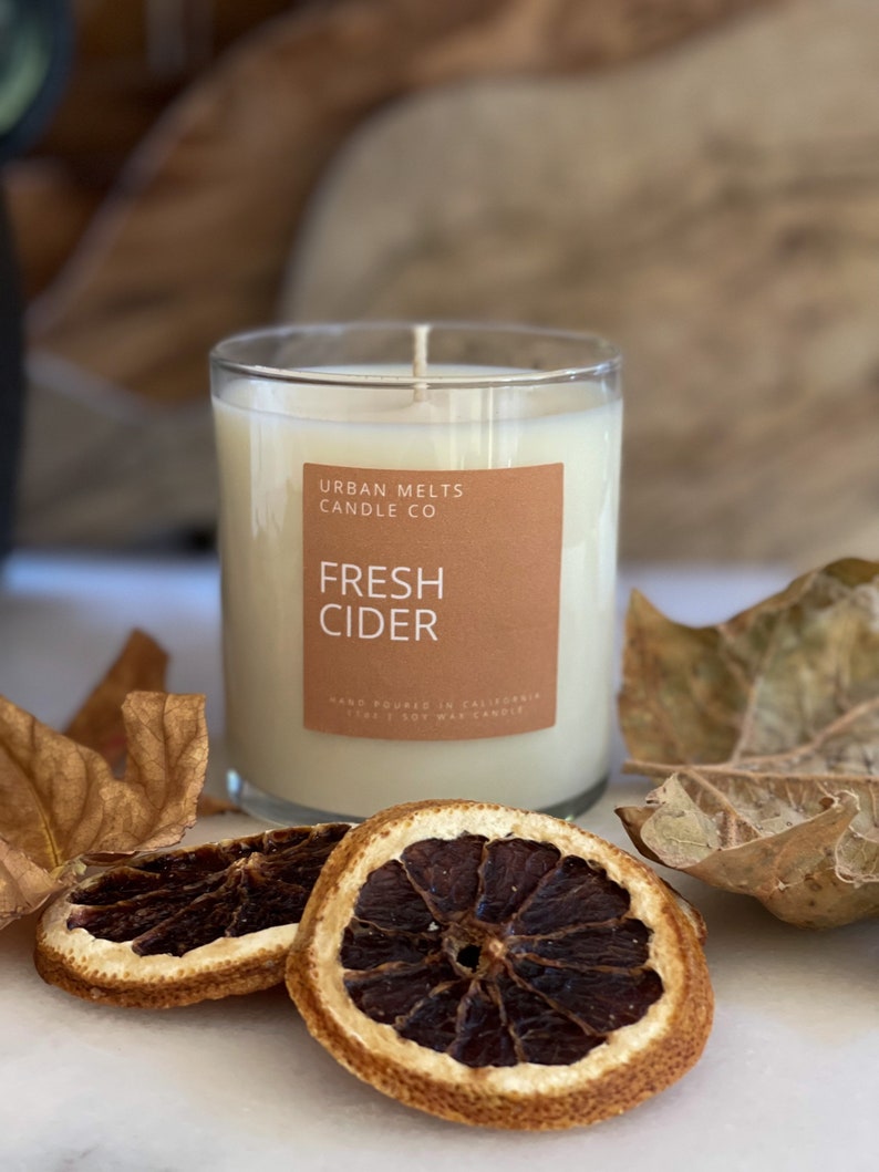 May include: A clear glass candle jar with a brown label that reads "Urban Melts Candle Co. Fresh Cider" and the text "Hand Poured in California 100% Soy Wax Candle". The candle is surrounded by dried orange slices and fall leaves.