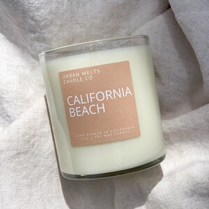 May include: A clear glass candle jar with a white soy wax candle inside. The label on the jar is brown and reads "Urban Melts Candle Co. California Beach Hand Poured in California 11oz Soy Wax Candle".