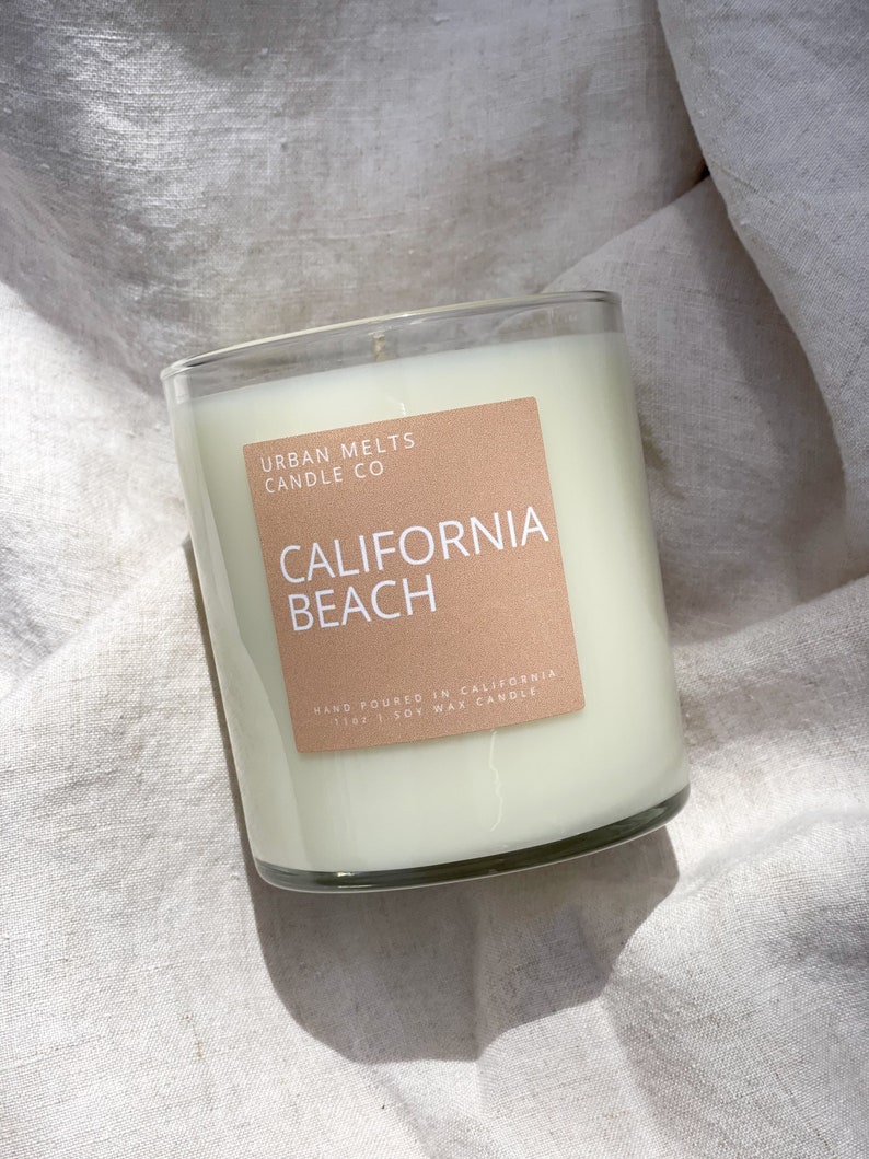 May include: A clear glass candle jar with a white soy wax candle inside. The label on the jar is brown and reads "Urban Melts Candle Co. California Beach Hand Poured in California 11oz Soy Wax Candle".