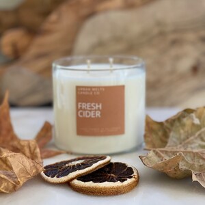 May include: A clear glass candle jar with a brown label that reads "Urban Wicks Candle Co. Fresh Cider". The candle is surrounded by dried orange slices and fall leaves on a white marble surface.