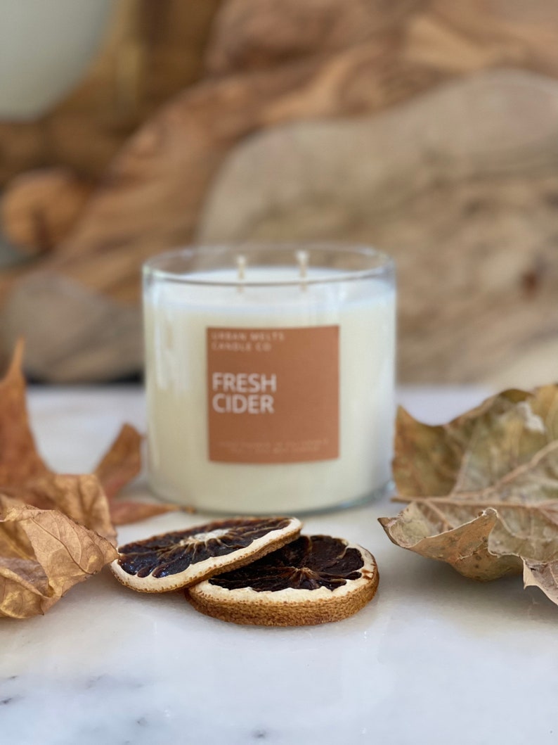 May include: A clear glass candle jar with a brown label that reads "Urban Wicks Candle Co. Fresh Cider". The candle is surrounded by dried orange slices and fall leaves on a white marble surface.