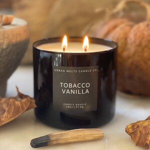 Puede incluir: Un tarro de vela de vidrio negro con una etiqueta blanca que dice "Urban Melts Candle Co. Tobacco Vanilla Candle Bougie 18oz | 510g". La vela est&aacute; encendida y tiene dos mechas.