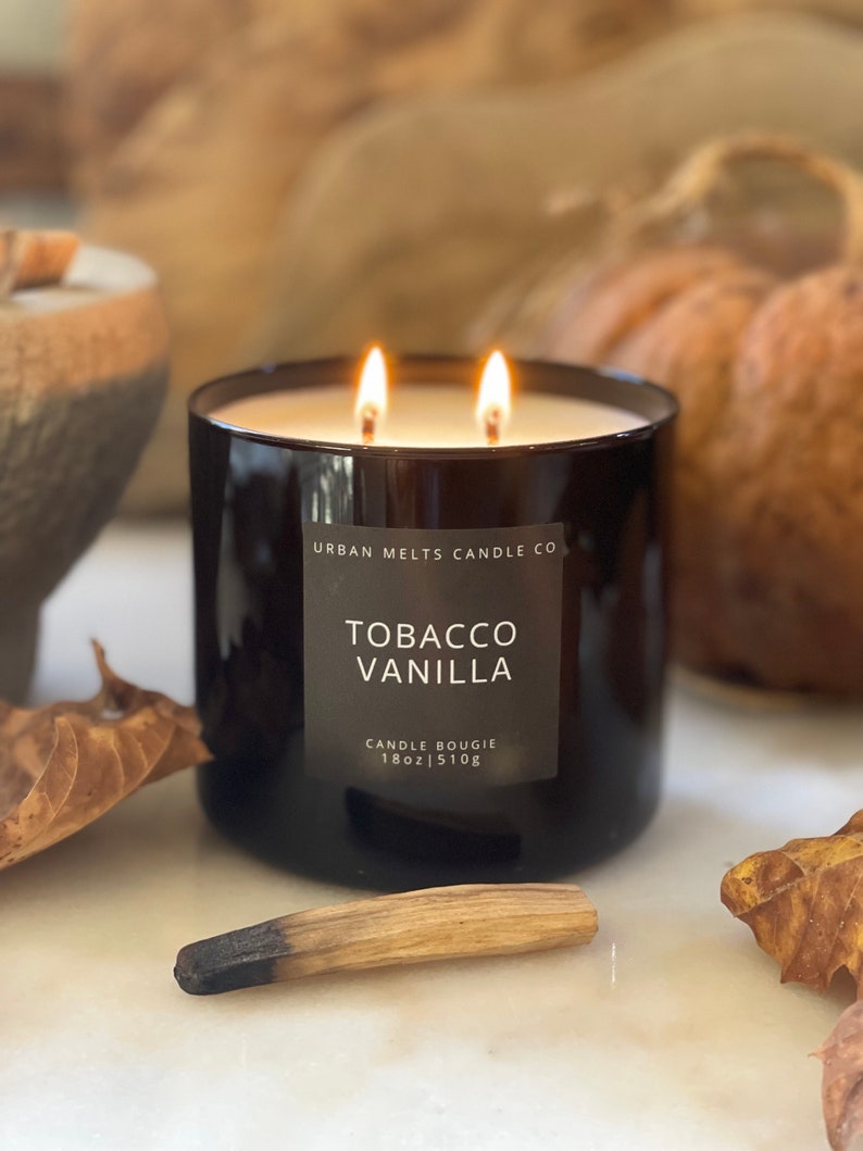 Puede incluir: Un tarro de vela de vidrio negro con una etiqueta blanca que dice "Urban Melts Candle Co. Tobacco Vanilla Candle Bougie 18oz | 510g". La vela est&aacute; encendida y tiene dos mechas.