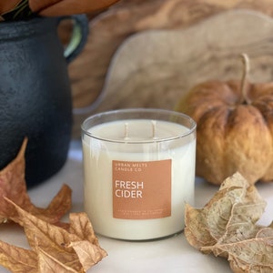 May include: A clear glass candle jar with a brown label that reads "Urban Melts Candle Co. Fresh Cider"  Hand poured in California. Soy Wax Candle. The candle is sitting on a white surface with fall leaves and a small pumpkin.