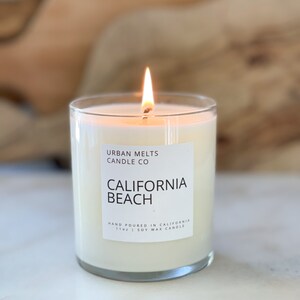 May include: A white candle with a clear glass jar and a white label that reads "URBAN MELTS CANDLE CO CALIFORNIA BEACH HAND POURED IN CALIFORNIA 110g | SOY WAX CANDLE". The candle is lit and the flame is visible.