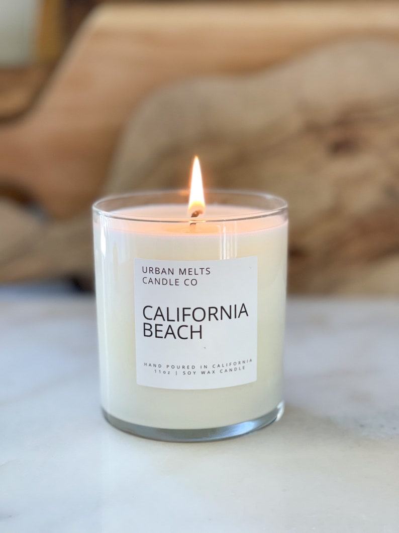 May include: A white candle with a clear glass jar and a white label that reads "URBAN MELTS CANDLE CO CALIFORNIA BEACH HAND POURED IN CALIFORNIA 110g | SOY WAX CANDLE". The candle is lit and the flame is visible.