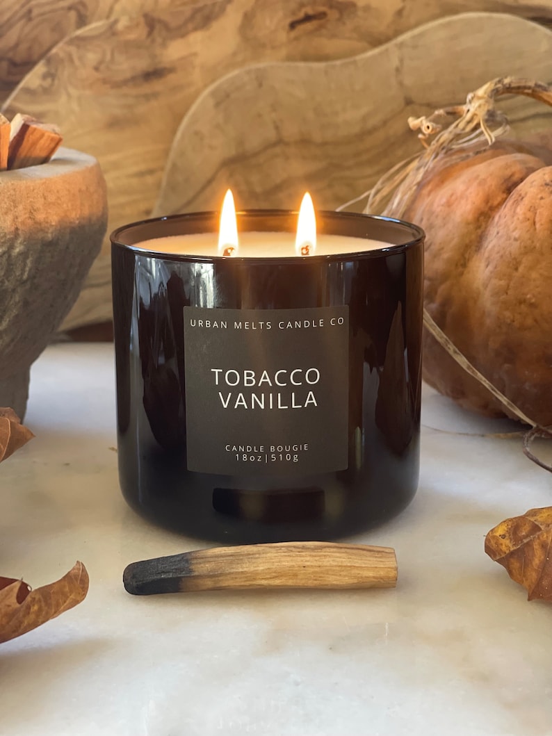 Puede incluir: Un tarro de vela de vidrio marr&oacute;n oscuro con dos mechas y una etiqueta que dice "Urban Melts Candle Co. Tobacco Vanilla Candle Bougie 18oz | 510g".