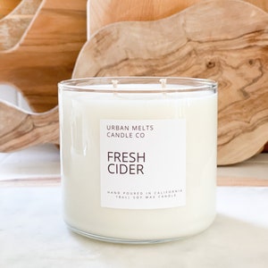 May include: A clear glass candle jar with a white label that reads "URBAN MELTS CANDLE CO" and "FRESH CIDER". The label also states "HAND POURED IN CALIFORNIA 18oz SOY WAX CANDLE".